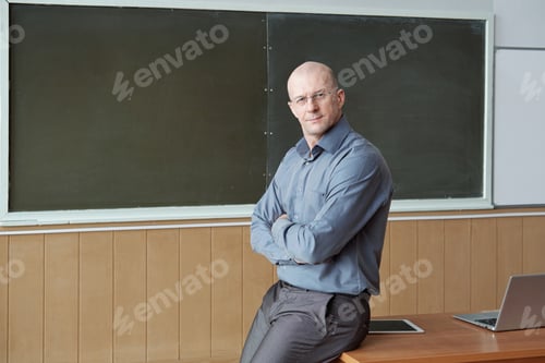 Preview: Bald professor of university in smart casualwear sitting on desk against blackboard