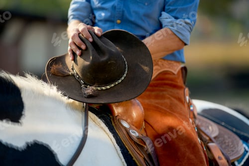 Preview: Cowboy in blue shirt adjusting a brown cowboy hat while riding a black and white horse.