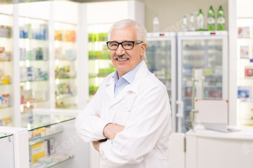 Preview: Aged drugstore worker in whitecoat and eyeglasses standing against displays