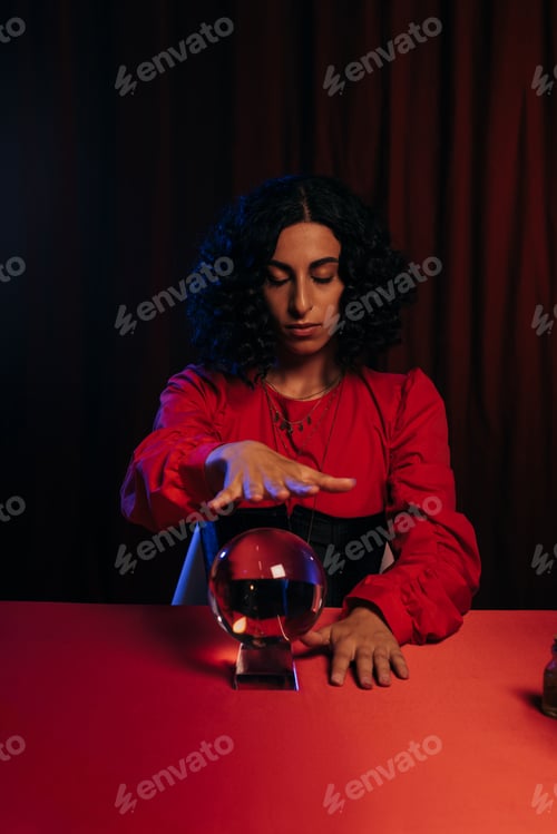 Preview: brunette fortune teller with closed eyes holding hand over crystal ball on dark background with red