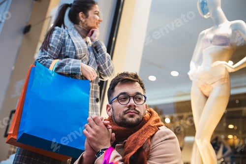 Preview: Man sitting nervously while a happy woman in shopping looking at the shop window.