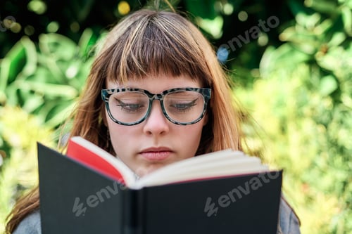 Preview: Close-up portrait of blue-eyed Caucasian blonde young woman with glasses reading a book in the park