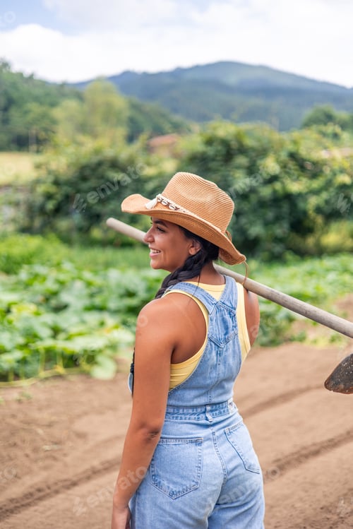 Preview: vertical Latin farmer woman carrying hoe on shoulder smiling in cultivated field