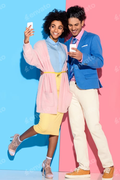 Preview: Young african amercian girl taking selfie and hugging stylish guy who looks at his phone on pink and