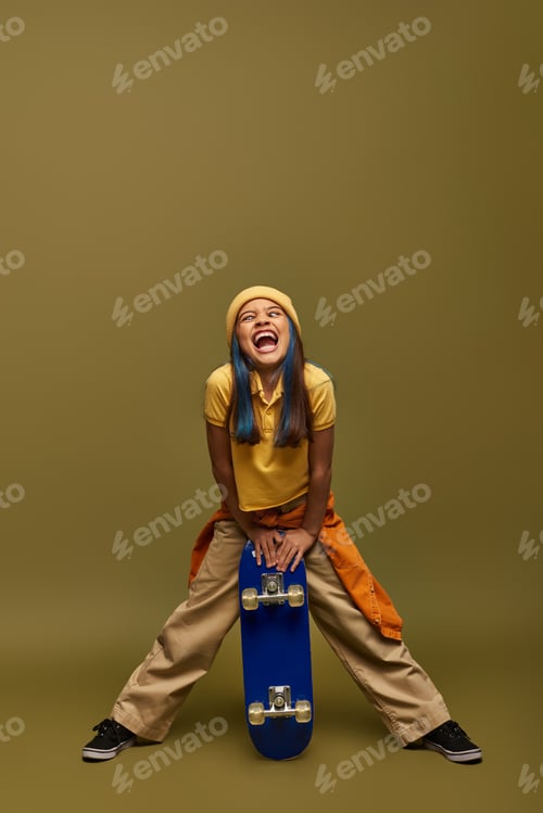 Preview: Full length of excited preteen child with colored hair wearing urban outfit and hat while grimacing
