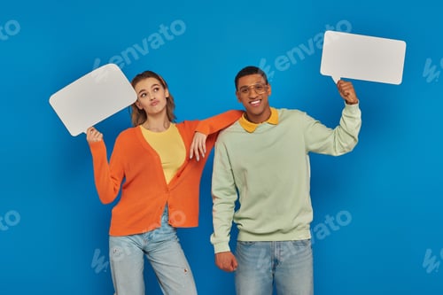 Preview: cheerful interracial couple in smart casual clothes holding blank speech bubbles on blue backdrop