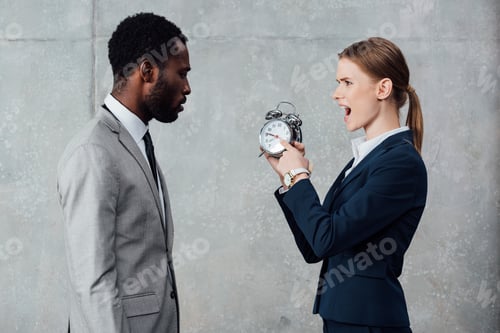 Preview: african american businessman looking at yelling businesswoman pointing at clock on grey background