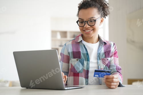 Preview: Mixed race teen girl holds bank credit card uses online banking services on laptop. E-banking