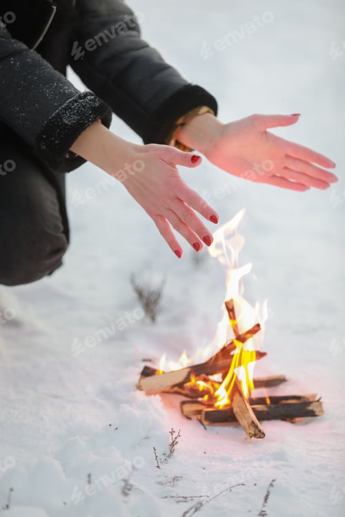 Preview: cropped photo of young woman warms her hands over bonfire in winter forest. Close-up
