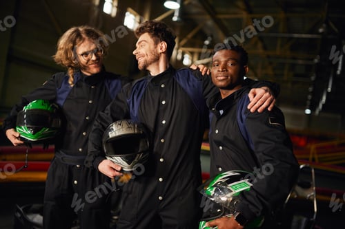 Preview: group of multicultural and happy go kart drivers in protective suits hugging and holding helmets