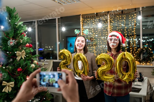 Preview: Two young happy businesswomen with inflatable numbers posing for colleague