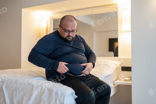 Preview: Man suffering from extra weight touching abdomen sitting on bed in bedroom.