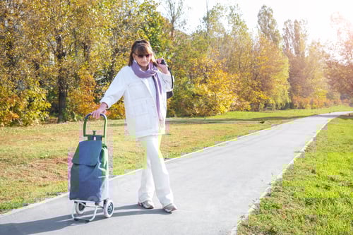 Vorschau: Stilvolle ältere kaukasische braunhaarige Frau mit Sonnenbrille und weißem Jeansanzug mit Einkaufstasche