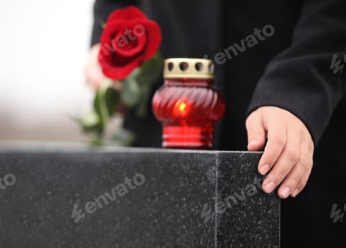 Preview: Woman holding red rose near black granite tombstone with candle outdoors, closeup. Funeral ceremony