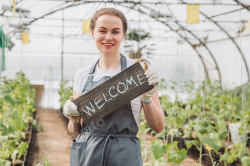 Preview: Female greenhouse worker holding welcome sign while standing by plants in greenhouse