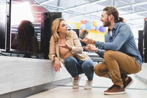 Preview: smiling boyfriend and girlfriend pointing with hands at new tv and talking in home appliance store