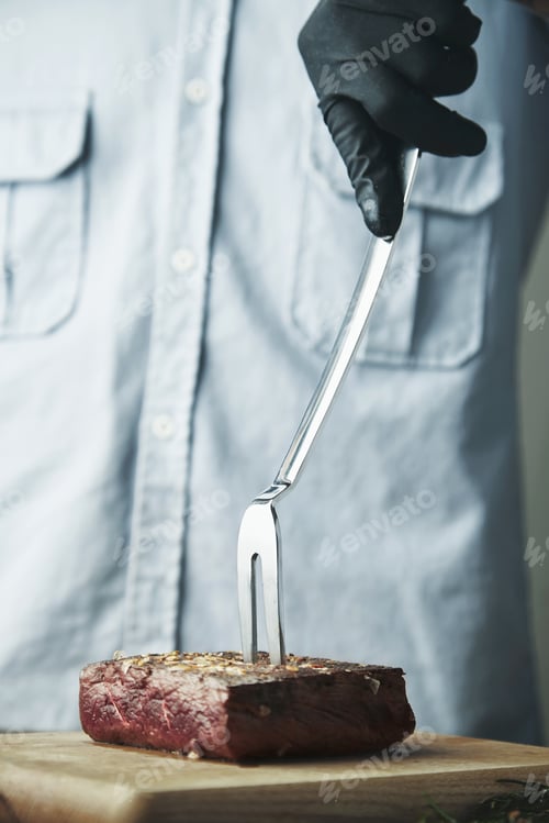 Preview: Hand in black glove holds steel fork with grilled meat
