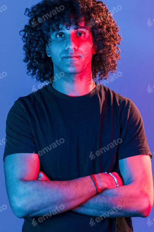 Preview: Posing for a camera. Young beautiful man with curly hair is indoors in the studio with neon lighting