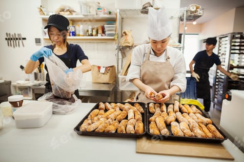 Preview: Two women and man working in a bakery, preparing freshly baked rolls on large trays.