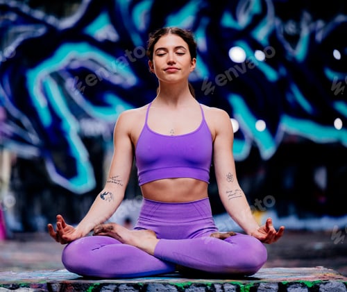 Preview: Woman meditating in a yoga pose wearing a purple outfit in front of vivid graffiti art. USA