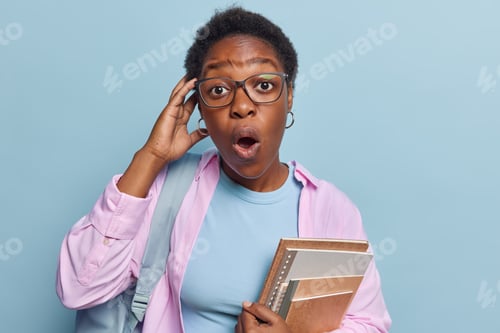 Preview: Horizontal shot of shocked dark skinned young woman with curly hair keeps hand on head has mouth