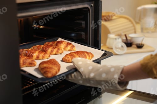 Preview: Woman taking baking sheet with tasty croissants out of oven indoors, closeup