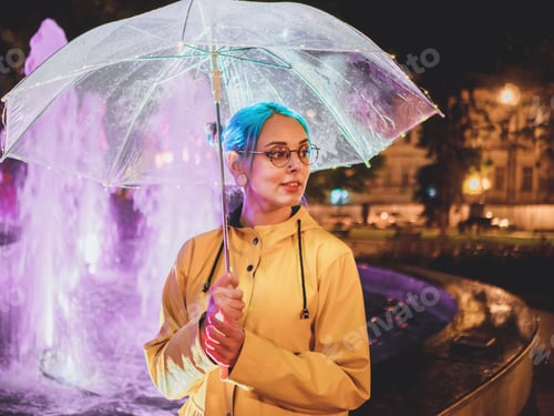 Preview: Young pretty girl with blue dyed hair in yellow raincoat and with transparent umbrella stands near