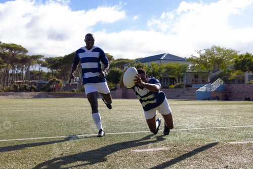 Preview: Two African American young male athletes in striped jerseys play rugby under the sun on a field
