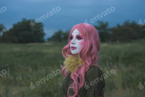 Preview: Young woman made up as a sad clown in a lonely field. Halloween makeup.