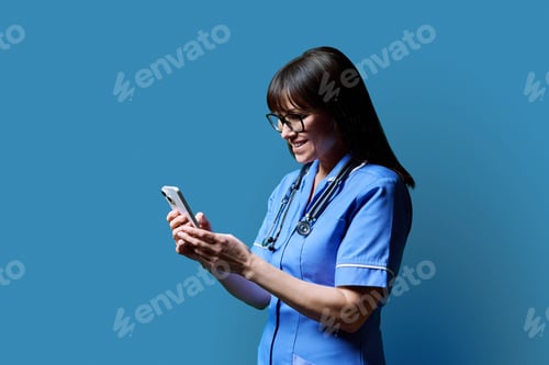 Preview: Female nurse using smartphone, profile view on blue studio background