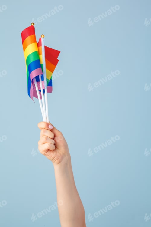 Preview: Closeup of a hand waving a small rainbow flag against a light blue background