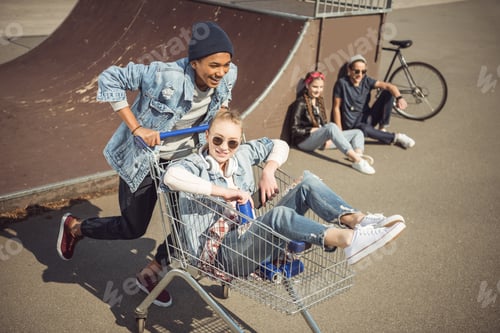 Preview: Boy carrying happy girl in shopping cart while friends sitting near ramp at skateboard park