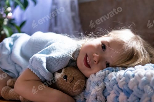 Preview: A little girl lies on a blanket and hugs her teddy bear. the girl looks at the camera