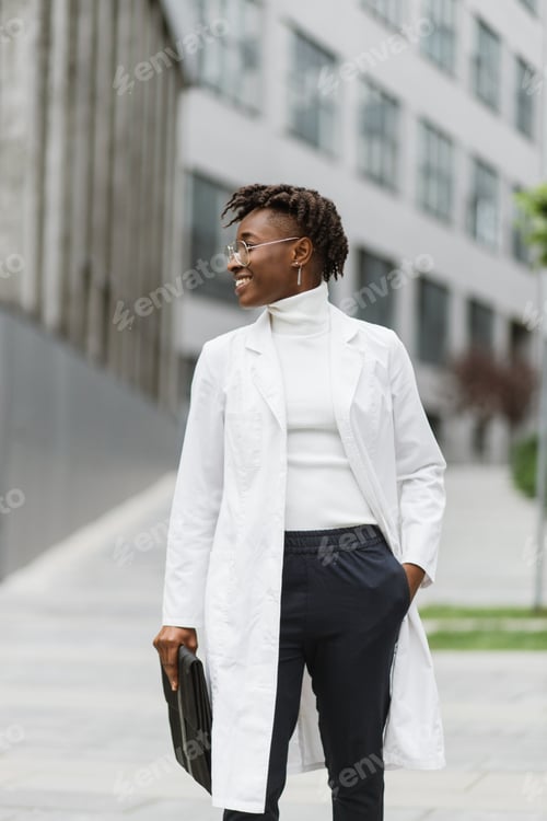 Preview: African American female doctor or student in white medical coat, standing outside modern clinic