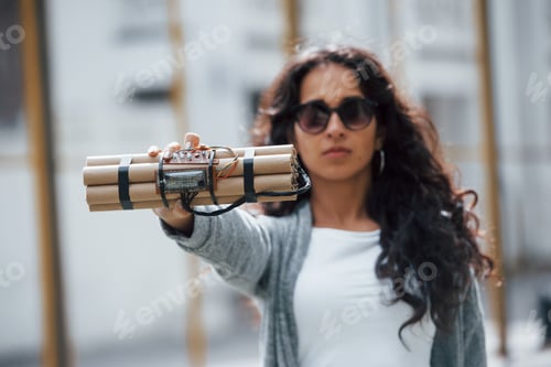 Preview: Serious girl in eyewear. Showing time bomb. Young woman holding dangerous explosive weapon in hand