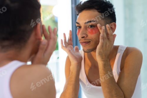 Preview: Biracial transgender man looking in mirror and applying under-eye patches in bathroom