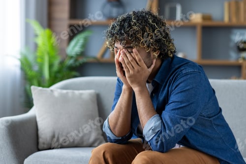 Preview: Desperate and depressed man sitting on sofa in living room at home, burying face in hands, feeling