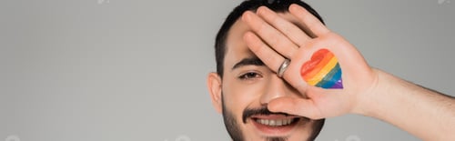 Preview: Cheerful homosexual man covering face with lgbt flag on hand isolated on grey, banner