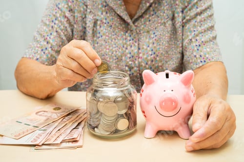 Preview: Asian elderly woman putting coin into pink piggy bank for saving money and insurance, poverty