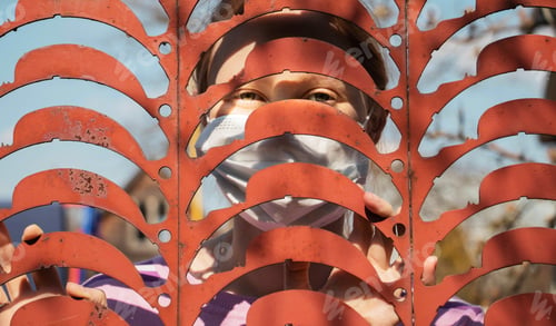 Preview: girl in a protective medical mask behind the iron bars of the fence