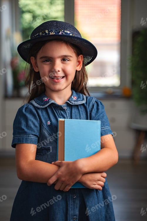 Preview: Smiling schoolgirl holding a book and wearing a hat