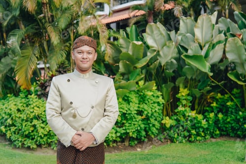Preview: A confident smiling Asian man wearing traditional Javanese beskap and batik standing in a garden.
