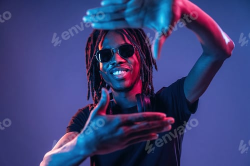 Preview: Gesture of capturing the photo. African American guy is studio with neon lighting
