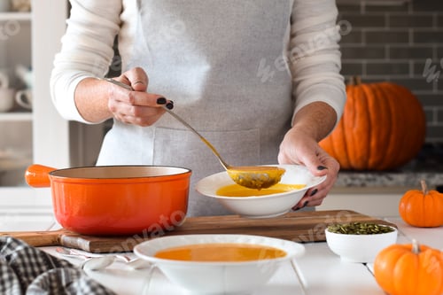 Preview: Woman using ladle to pour pumpkin soup into bowls on kitchen counter table