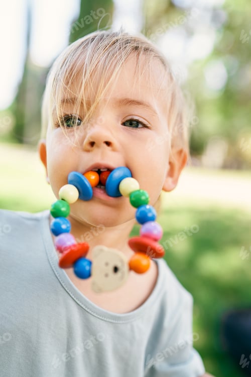 Preview: Little girl with a toy bracelet in her mouth on a green lawn. Portrait