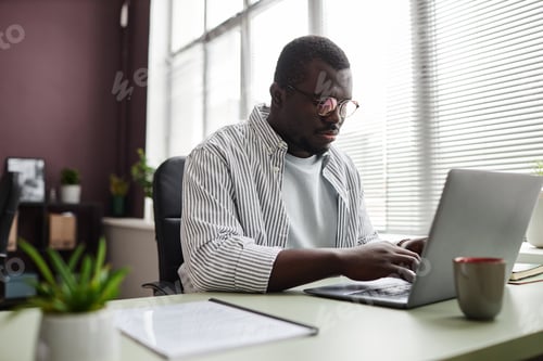 Preview: Focused black man using laptop working at desk in office wearing casual clothes