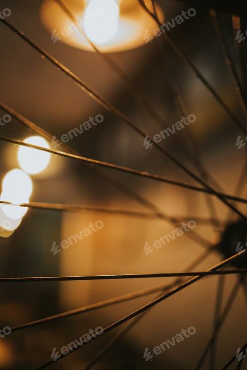 Preview: Selective focus shot of bicycle spokes in a dimly lit room with lightbulbs in the blurred background