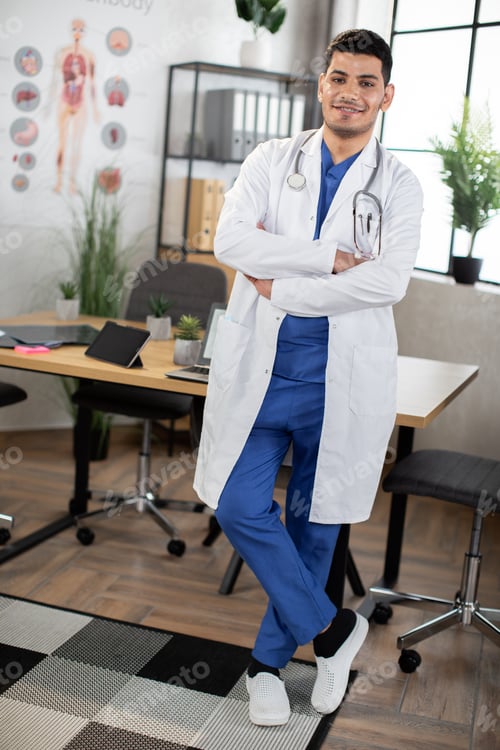 Preview: Arabic Indian medical doctor, wearing blue scrubs and white gown standing with arms crossed