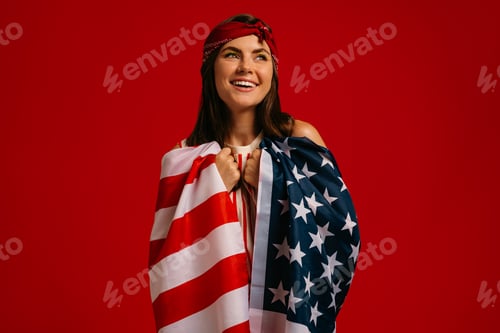 Preview: Attractive young hipster woman carrying American flag on shoulders and smiling on red background