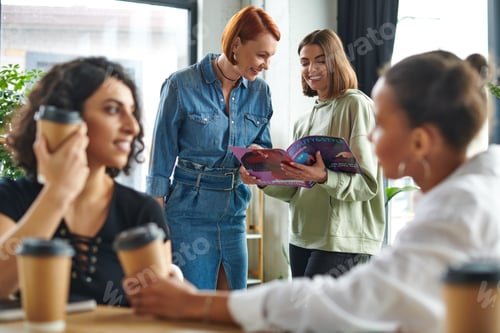 Preview: cheerful girlfriends reading magazine while multiethnic women drinking coffee from paper cups